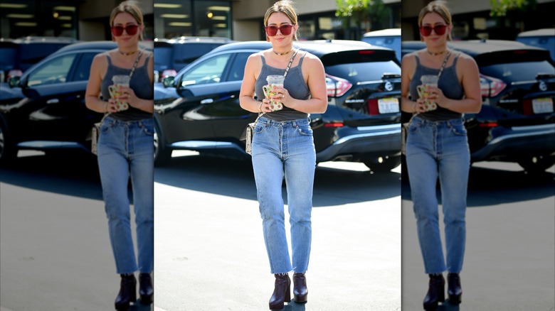 Hilary Duff photographed on the street holding a cup and wearing a gray tank top, jeans, black boots, and red sunglasses.