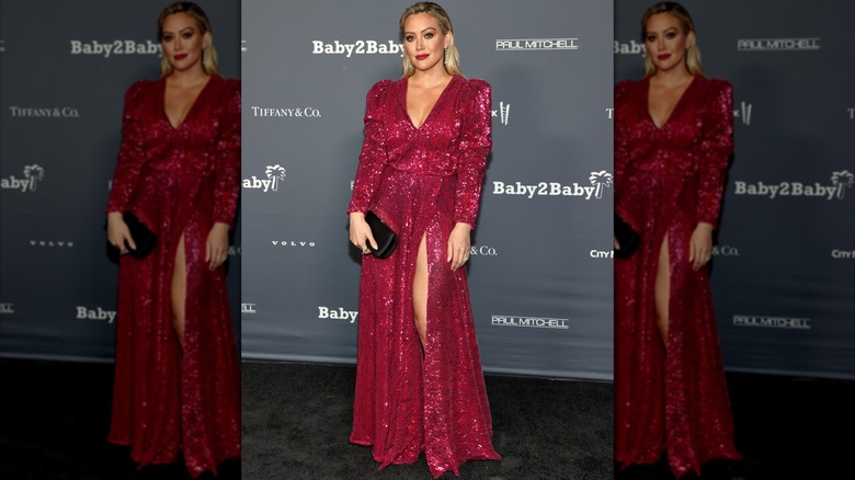 Hilary Duff at a red carpet event posing in a sparkly red dress with a deep-V neckline and slit in the skirt.