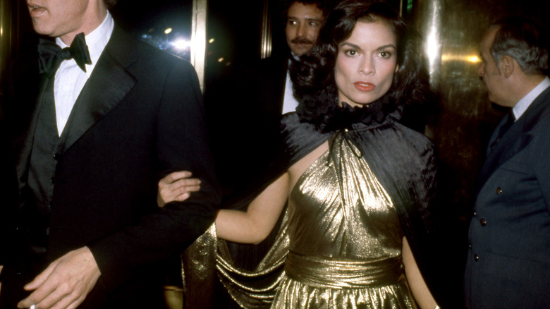 Bianca Jagger wearing a gold gown and black cape at a nighttime event.
