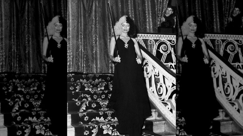 Bianca Jagger walking down ornate stairs wearing a black gown with a cane and split black-and-white hair.