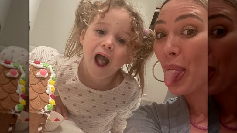 Hilary Duff and daughter Banks stick out their tongues while decorating a gingerbread house in their pajamas.
