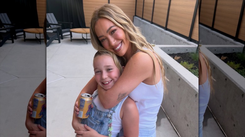 Hilary Duff wears her blond hair down in mermaid waves while wearing a white tank top and posing with daughter Banks during her "Lucky Me" tour in 2026.