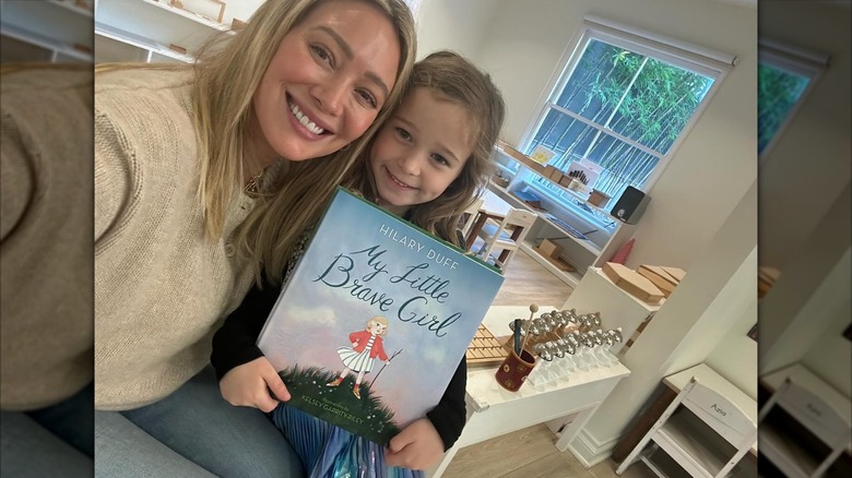Hilary Duff wears her blond hair straight down in a beige sweater and jeans while posing with her daughter Banks and her new book.