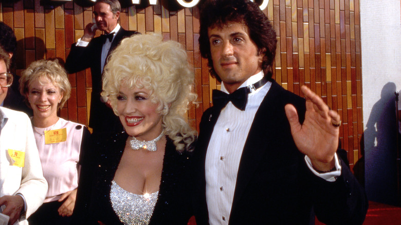 Dolly Parton in 1984 posing with Sylvester Stallone wearing a sparkly silver top with matching bow tie and a black sparkly jacket.