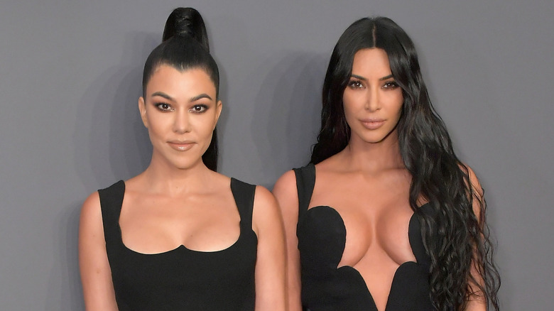 Kourtney Kardashian Barker and Kim Kardashian pose together wearing black against a gray background.