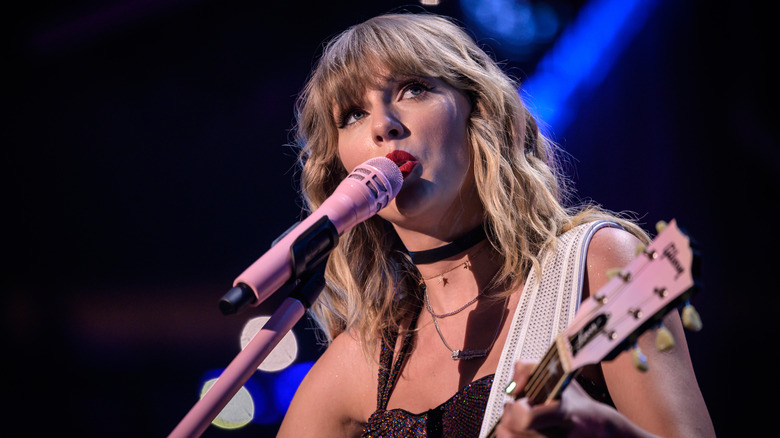 Taylor Swift wears bright red lipstick as she sings into a pink microphone and plays the guitar on stage.