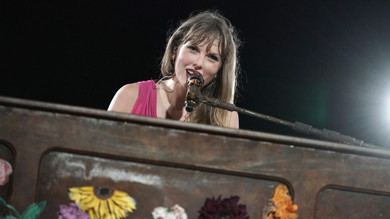 Taylor Swift smiles toward the camera while sitting at a piano and singing into a microphone.