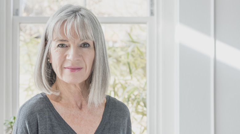 A woman with a gray bob with bangs standing in front of a window.