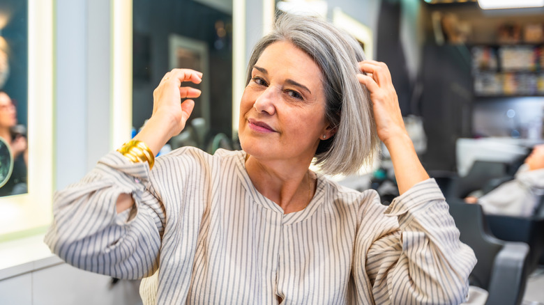 A woman with a chin-length gray bob haircut touching her hair in a salon.