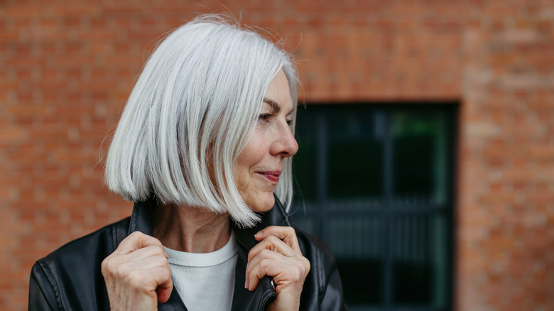 A woman with a blunt gray bob haircut looking to the side with her hands on the collar of her leather jacket.