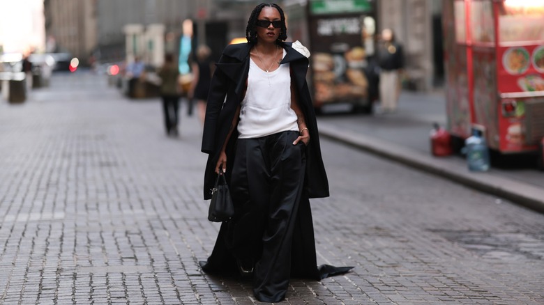 A woman walks down the street in black satin trousers, a white tank, and a black wool coat.