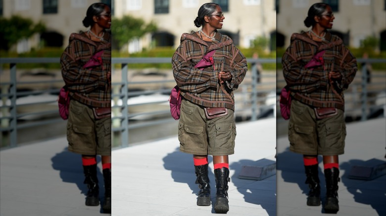 A woman wearing a brown and olive tartan jacket, olive cargo shorts, and black boots.