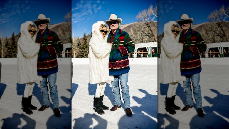 Goldie Hawn wearing a white fur coat links arms with Kurt Russell wearing a multi-colored sweater and jeans