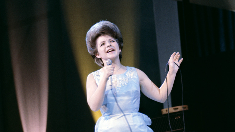 Brenda Lee wearing a blue dress with a bouffant hairdo singing into a microphone on stage in the '60s.