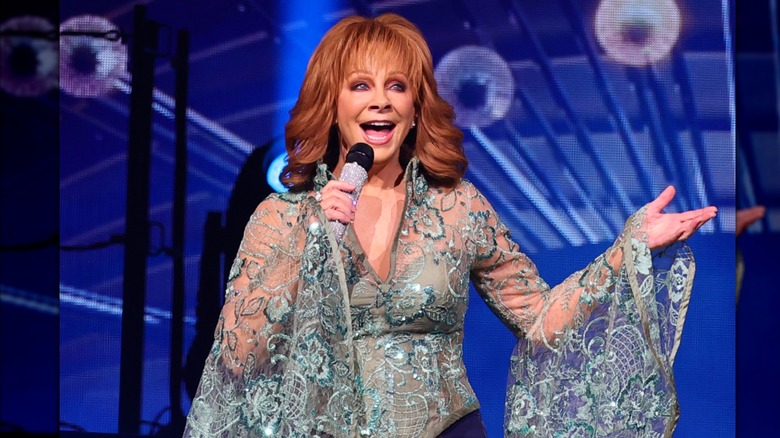 Reba McEntire wears a detailed turquoise top while singing into a bedazzled microphone on stage