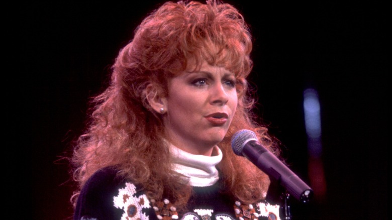 Reba McEntire sings into a microphone while sporting a voluminous curly hairstyle
