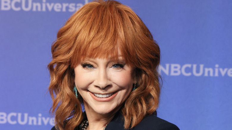 Reba McEntire smiles in front of a blue backdrop while posing on the red carpet