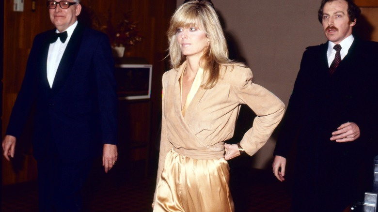Farrah Fawcett wears a beige jacket and gold jumpsuit