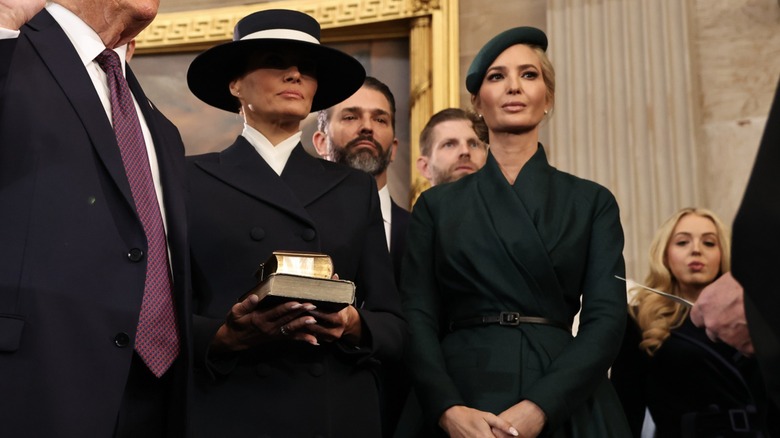Melania is wearing a navy blue coat with shoulder pads, a high-necked white blouse, and a wide-brimmed navy hat with a white ribbon. Her eyes are covered by the hat. Ivanka is wearing a dark green wrap coat with a matching skirt, and a green beret tilted to the side.