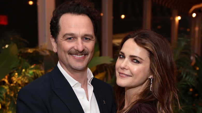 Matthew Rhys and Keri Russell pose together in front of a dimly-lit greenery background.