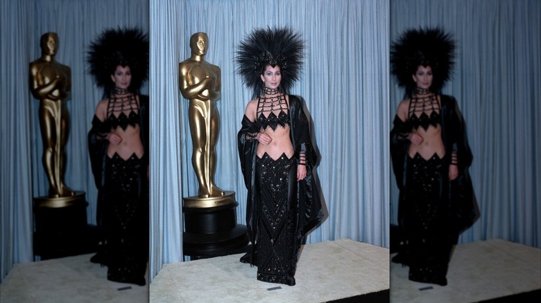 Cher wears a black sparkly two-piece set and feathered headdress to the 1986 Academy Awards