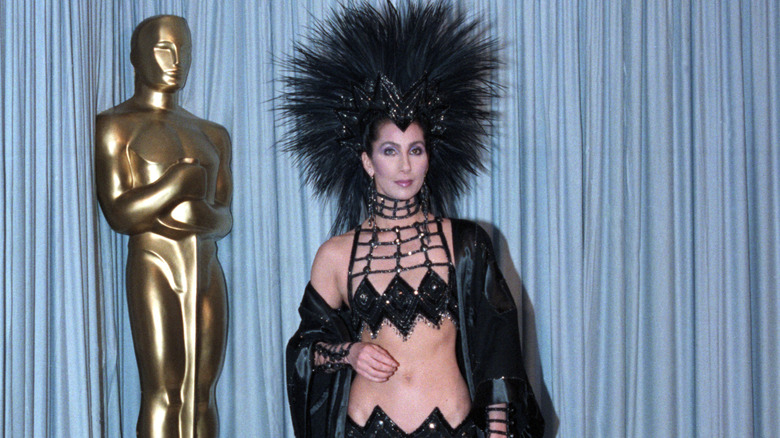 Cher wears a black sparkly two-piece set and feathered headdress to the 1986 Academy Awards