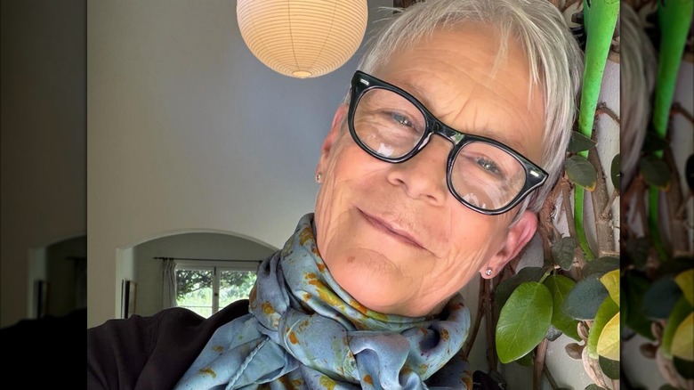 Jamie Lee Curtis tilting her head to the side while wearing glasses and a blue scarf in a selfie.