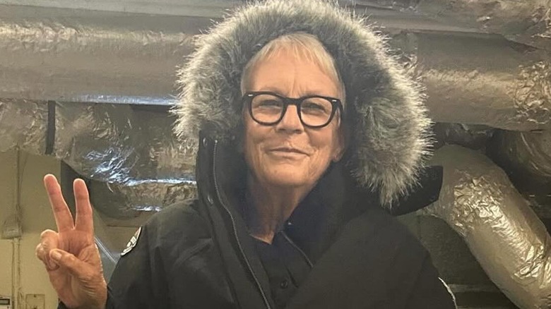 Jamie Lee Curtis giving a peace sign while wearing a hooded coat with fur trim.