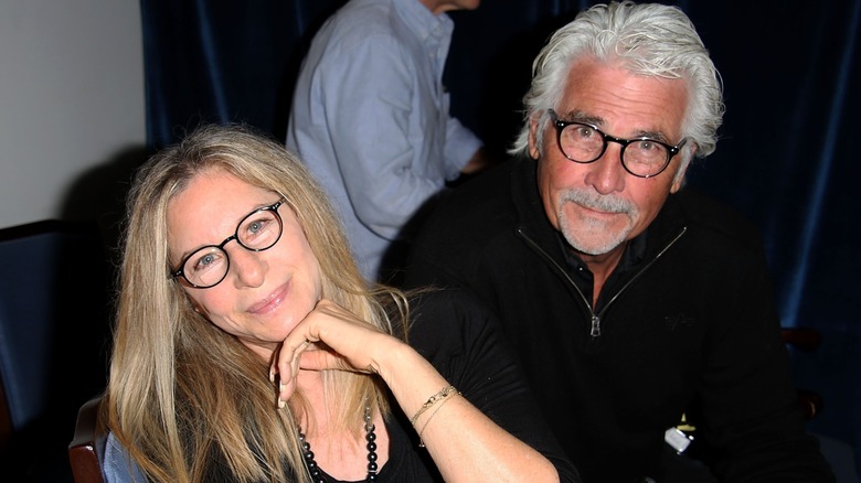 Barbra Streisand wearing glasses posing with her chin on her hand next to husband James Brolin wearing a black zip-up sweater and glasses.