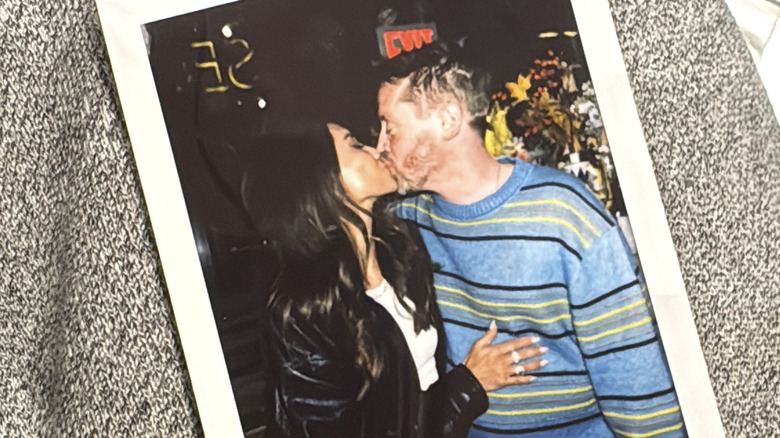 A photo of a Polaroid with Brenda Song in a leather jacket kissing Macaulay Culkin in a striped sweater.