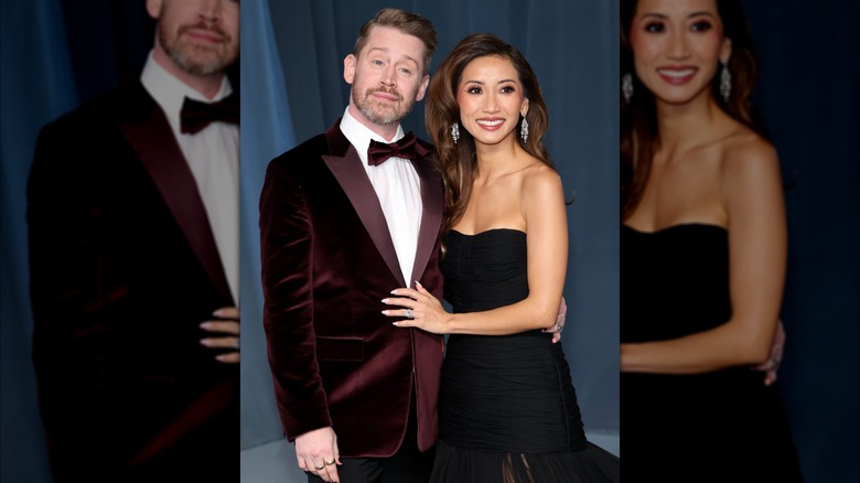 Macaulay Culkin in a burgundy suede Paul Smith suit posing next to Brenda Song in a SS23 Carolina Herrera gown.