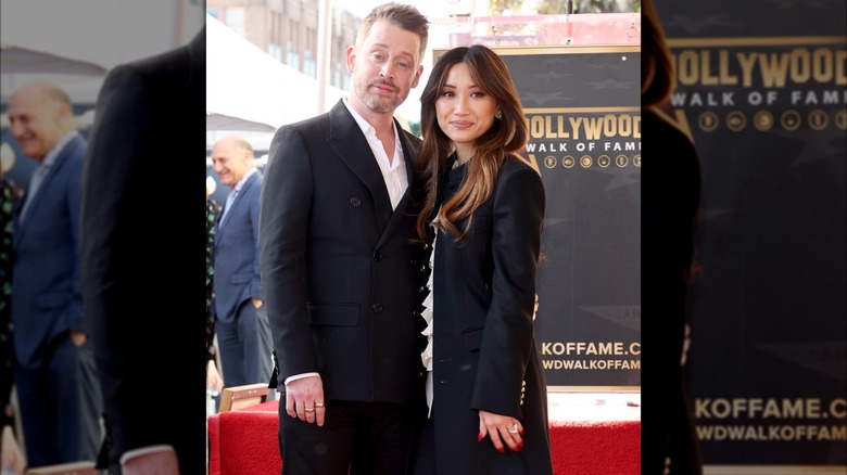 Macaulay Culkin in a black suit standing next to Brenda Song in a long black trench coat.
