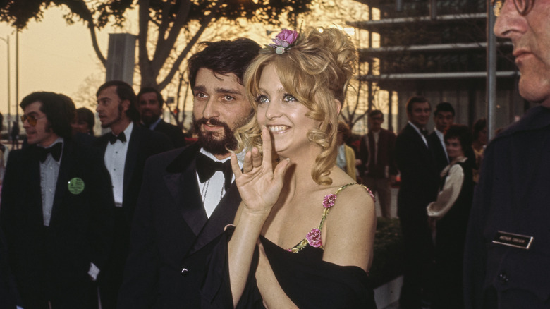 Goldie Hawn wears a flower in her hair and a black dress with floral straps in 1973