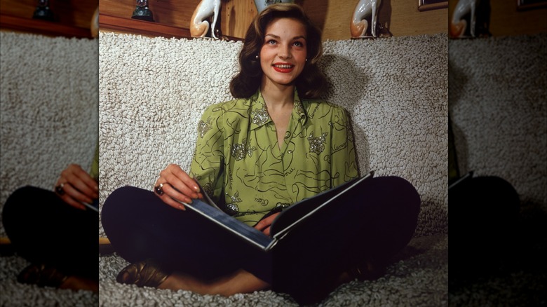Lauren Bacall sitting cross-legged with an open book in her lap wearing a green embellished blouse and red lipstick circa 1945