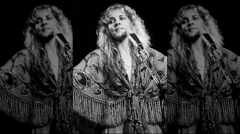 Black and white photo of Stevie Nicks in front of a microphone wearing a paisley fringe top performing with Fleetwood Mac in 1978.
