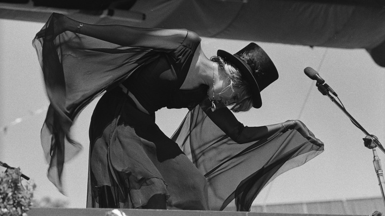 Black and white photo of Stevie Nicks wearing a flowing black outfit with a black top hat on stage performing with Fleetwood Mac in 1977.