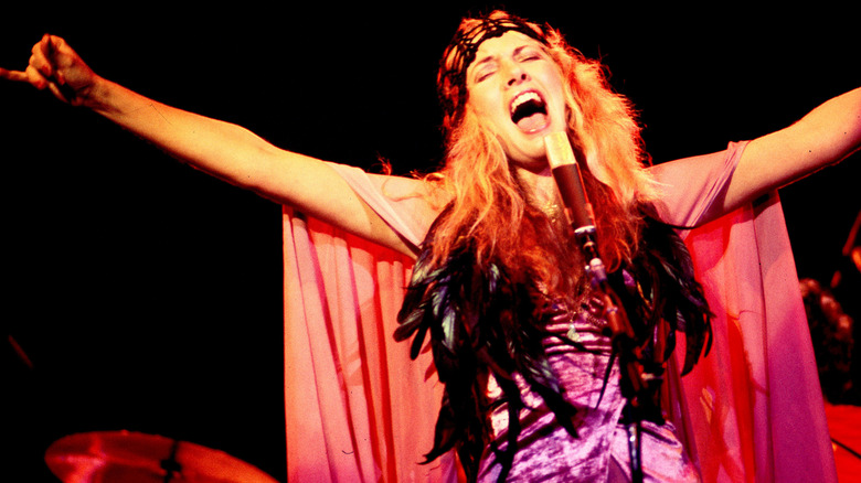 Stevie Nicks with her arms outstretched singing wearing a flowy cape and feathery outfit performing with Fleetwood Mac in 1978.