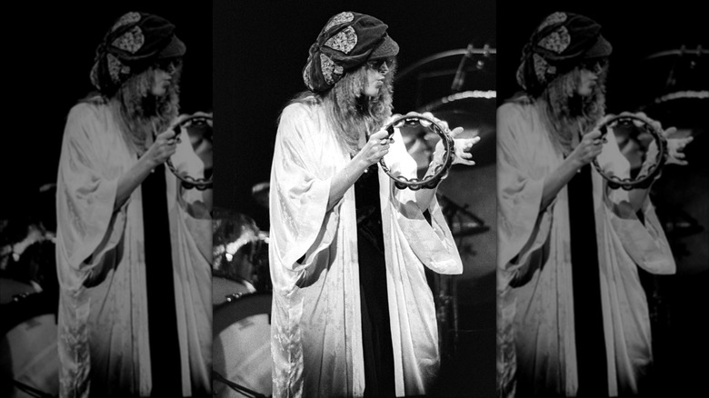 Black and white photo of Stevie Nicks wearing a floppy hat and a white robe playing a tambourine performing with Fleetwood Mac in 1977.