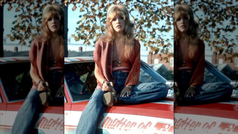 Stevie Nicks posing on a car wearing jeans and a red top with platform shoes and curtain bangs.