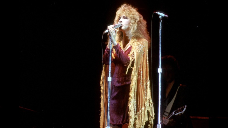 Stevie Nicks wearing a red outfit with a gold cape singing into a microphone performing with Fleetwood Mac in 1977.