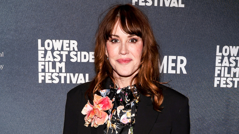 Molly Ringwald posing at an event wearing a black floral top and a black jacket.