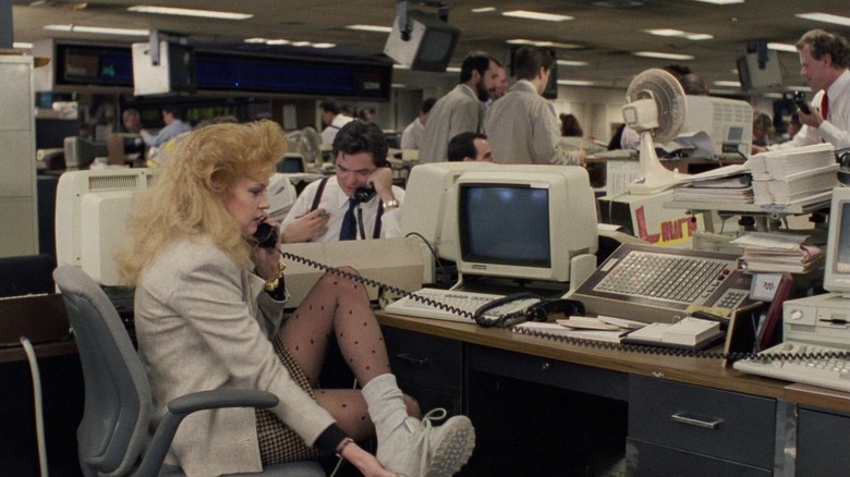 Melanie Griffith sitting at a desk wearing a beige blazer, plaid skirt, polka-dot tights, and white sneaker in "Working Girl."