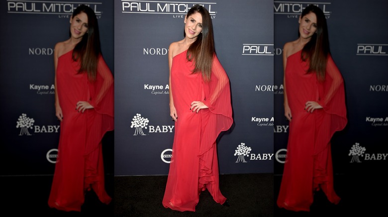 Soleil Moon Frye posing at a red carpet event wearing a red one-shoulder dress with a sheer flowy sleeve.