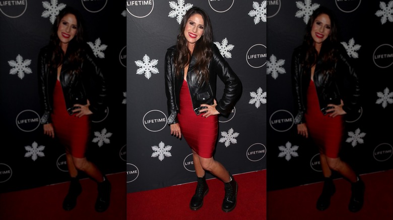 Soleil Moon Frye posing at a red carpet event wearing a red bandage dress with a black leather jacket and black boots.
