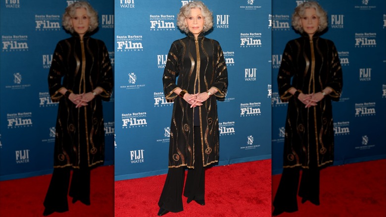 Jane Fonda posing at a red carpet event wearing flowing black pants and a black-and-gold tunic top.