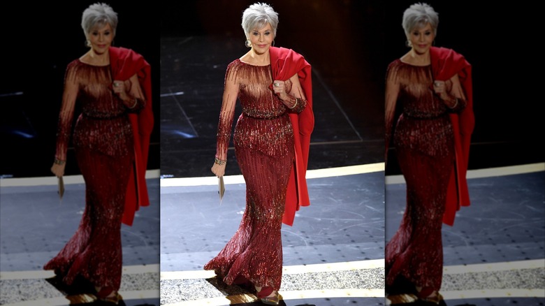 Jane Fonda posing at the Academy Awards wearing a glittery red dress with a bright red coat over her shoulder.
