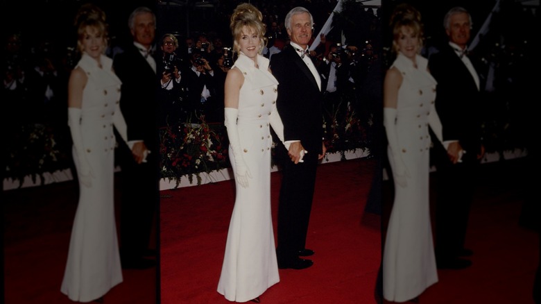 Jane Fonda posing on the Academy Awards red carpet wearing a white dress with gold buttons and long white gloves.