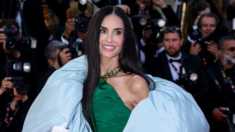 Demi Moore wears her black hair straight down with a middle part and an emerald-green high-neck gown and a puffy light blue jacket while attending the 79th Annual Cannes Film Festival in Cannes, France in 2026.