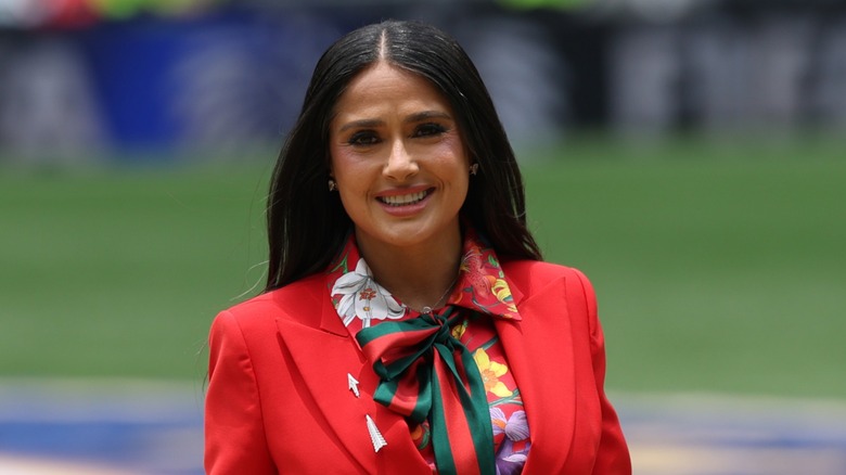 Salma Hayek wears her black hair straight down in a middle part along with a bright red suit jacket and a floral collared blouse underneath while speaking in the Opening Ceremony before a FIFA World Cup 2026 game in Mexico City.