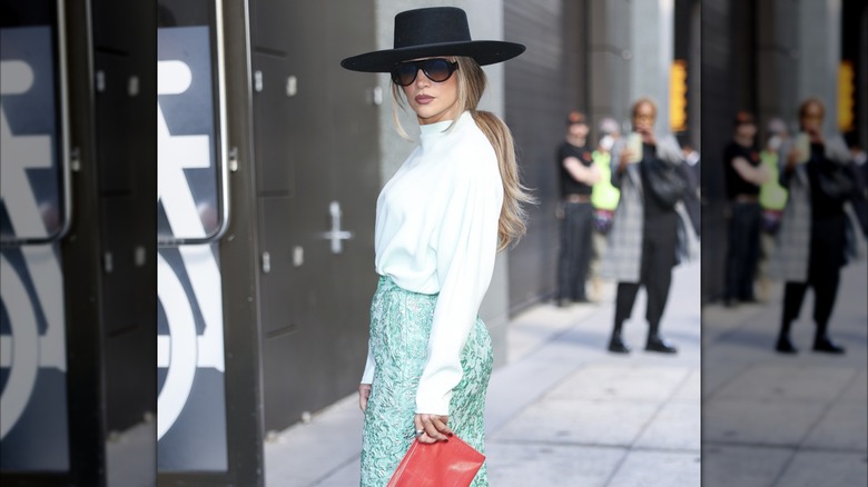 Jennifer Lopez posing outside in NYC wearing a large-brimmed black hat with sunglasses, a white top and green skirt.
